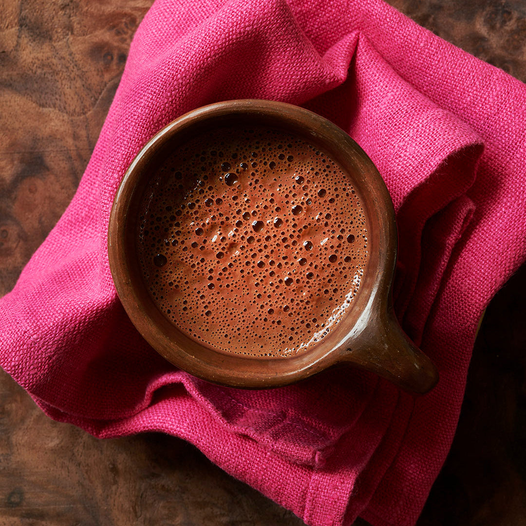 Ceremonial grade cacao from the Peruvian Andes in a handmade brown cup on a pink cotton napkin sitting on a dark wooden table, shot from above.