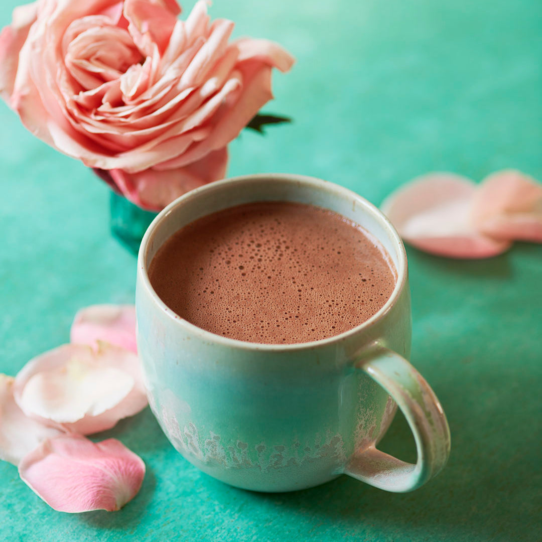 Ceremonial grade cacao from the Peruvian Andes in a handmade turquoise and pink ceramic mug surrounded by pink rose petals on a green background.